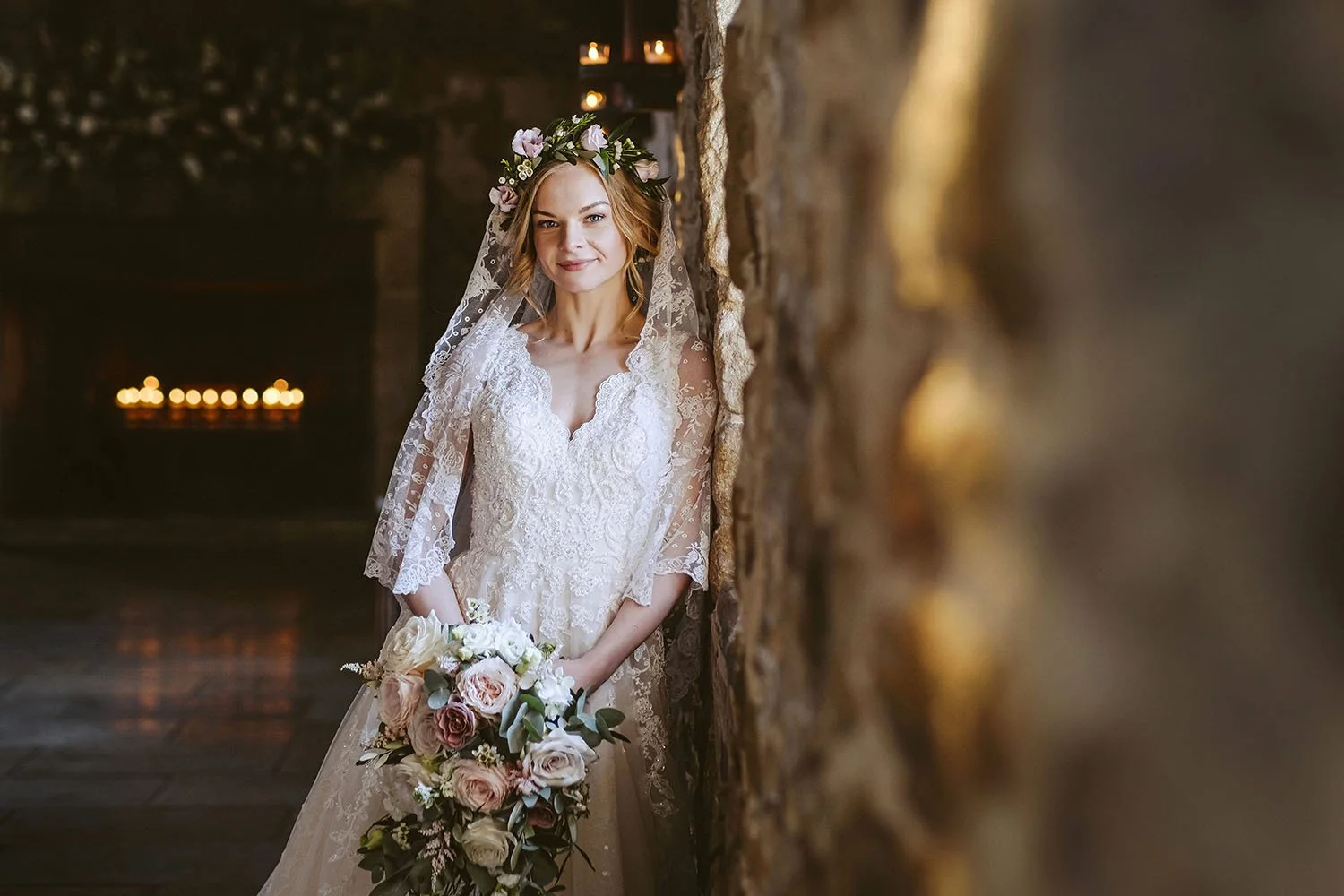 Healey Barn wedding: bride in a flower crown and lace veil holding a bouquet, leaning against a stone wall in a candlelit room.