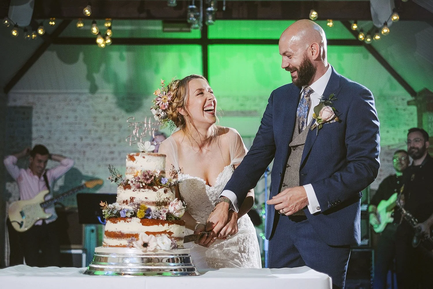 Shotton Grange wedding: bride and groom laughing as they cut a floral wedding cake while a band plays behind.