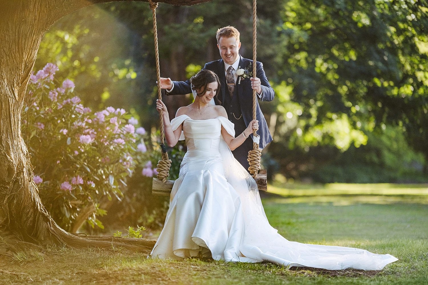Beamish Hall wedding: bride in a satin gown on a rope swing as the groom pushes her, both laughing