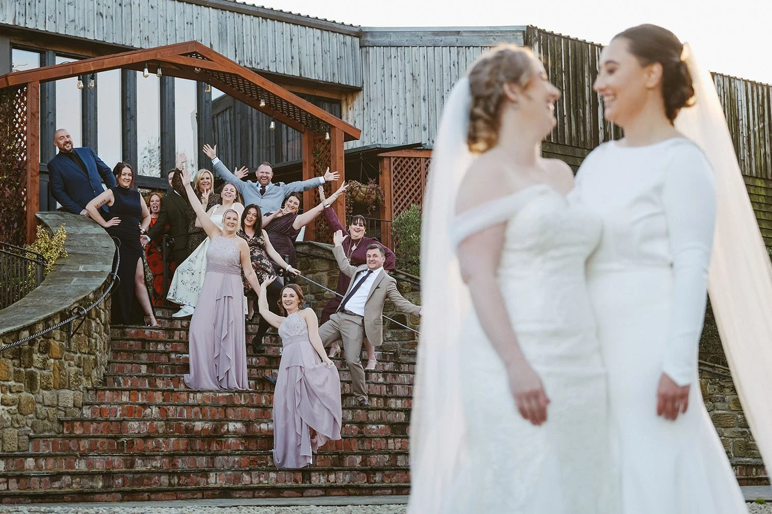 South Causey Inn wedding: two brides in veils smiling at each other in the foreground while the wedding party poses playfully