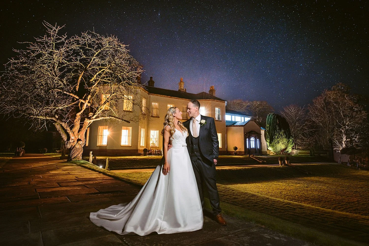 Lartington Hall wedding: bride and groom embracing at night beneath a starry sky, a lit country house and a bare tree behind them.