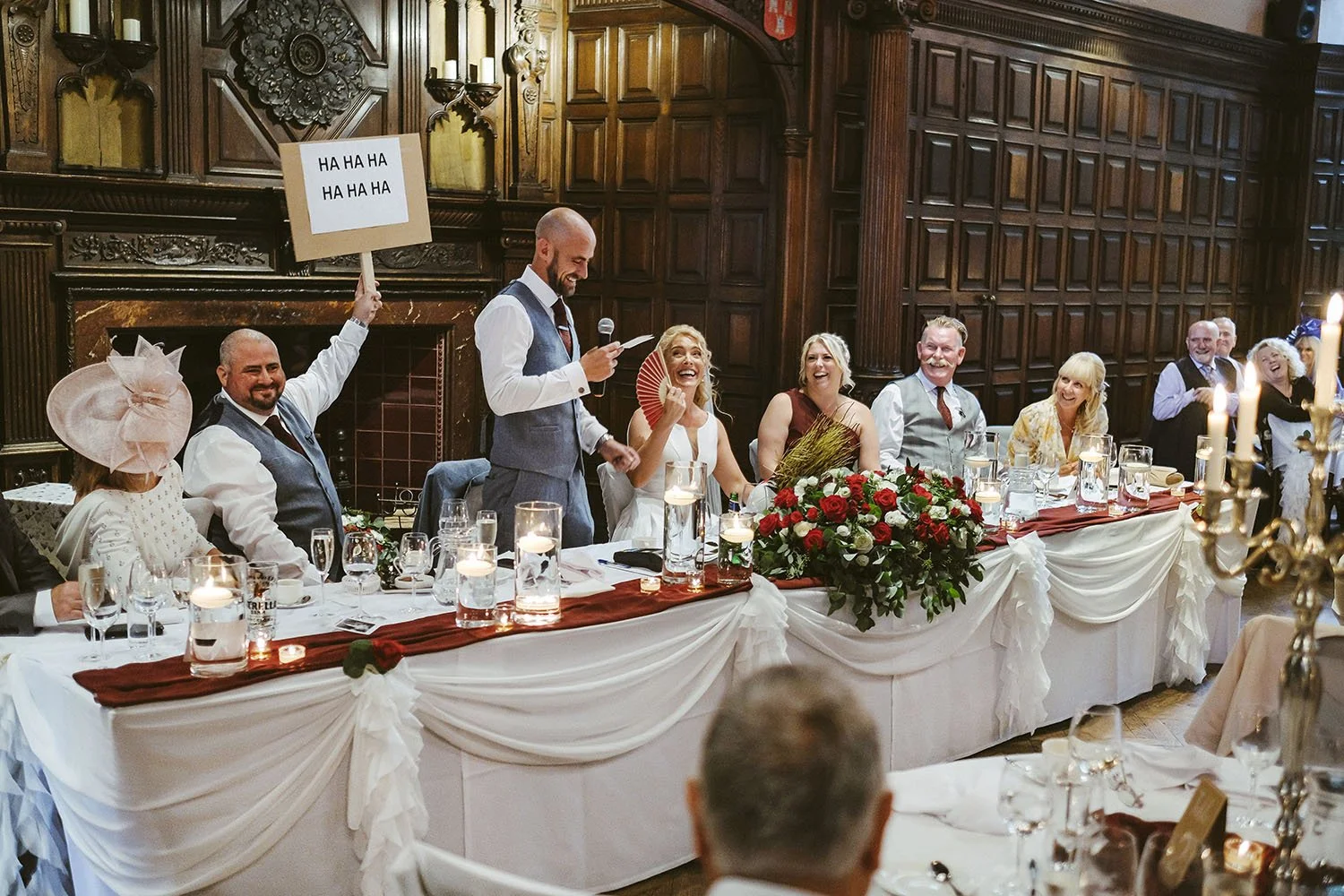 Jesmond Dene House wedding: best man giving a speech at a long top table in an oak-panelled hall as guests laugh.