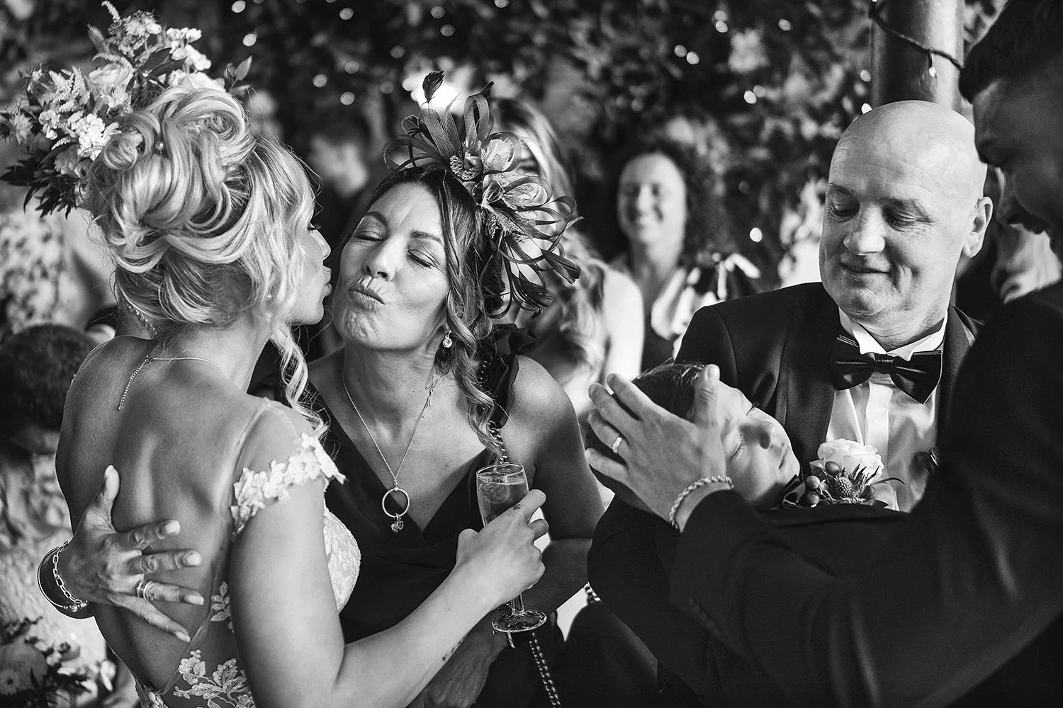 South Causey Inn wedding: bride in a flower crown being kissed on the cheek by a guest in a fascinator, in black and white.