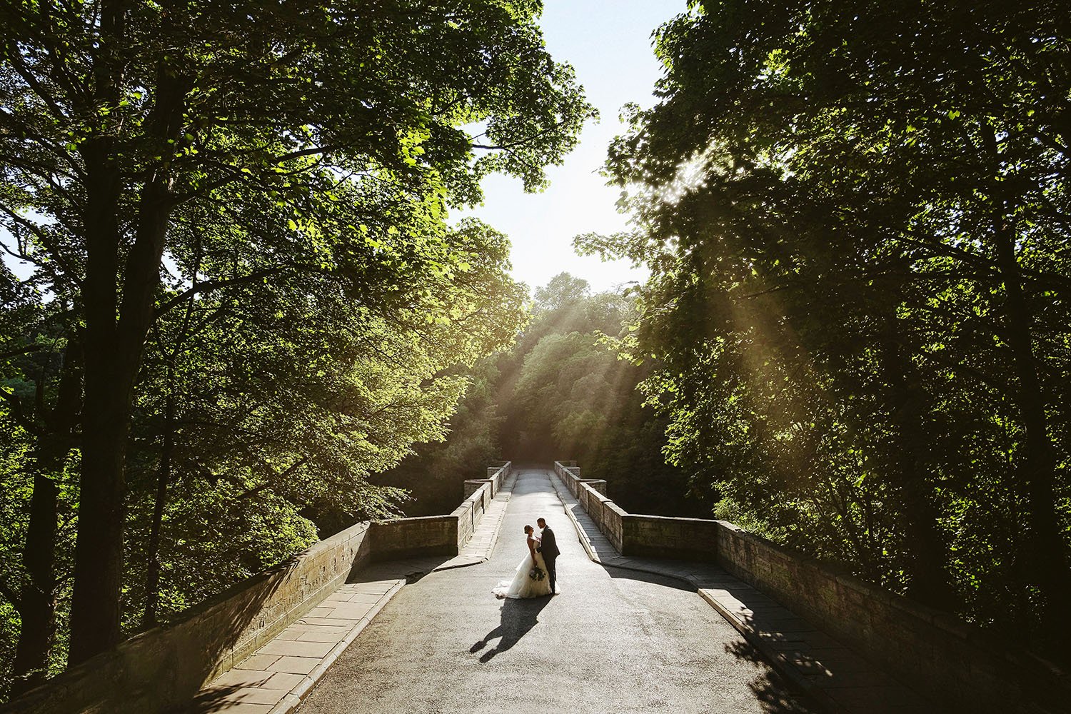 Durham Castle wedding: bride and groom walking together on a stone bridge through sunlit trees.