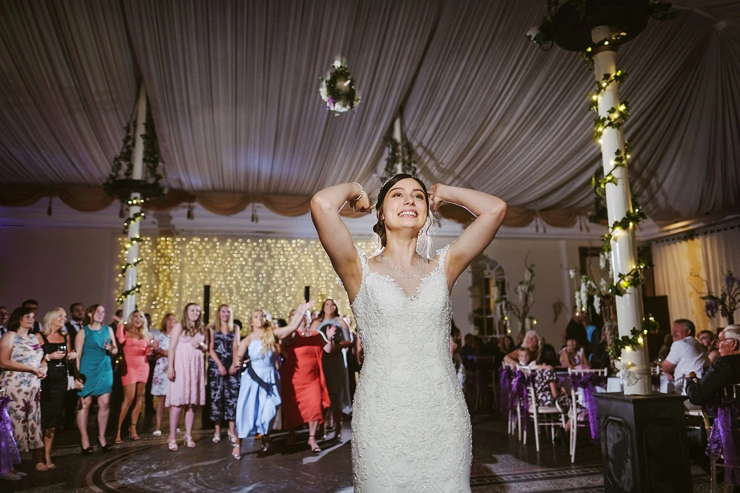 Beamish Hall wedding: bride flexing her arms before the bouquet toss as guests wait in a ballroom with a fairy-light curtain