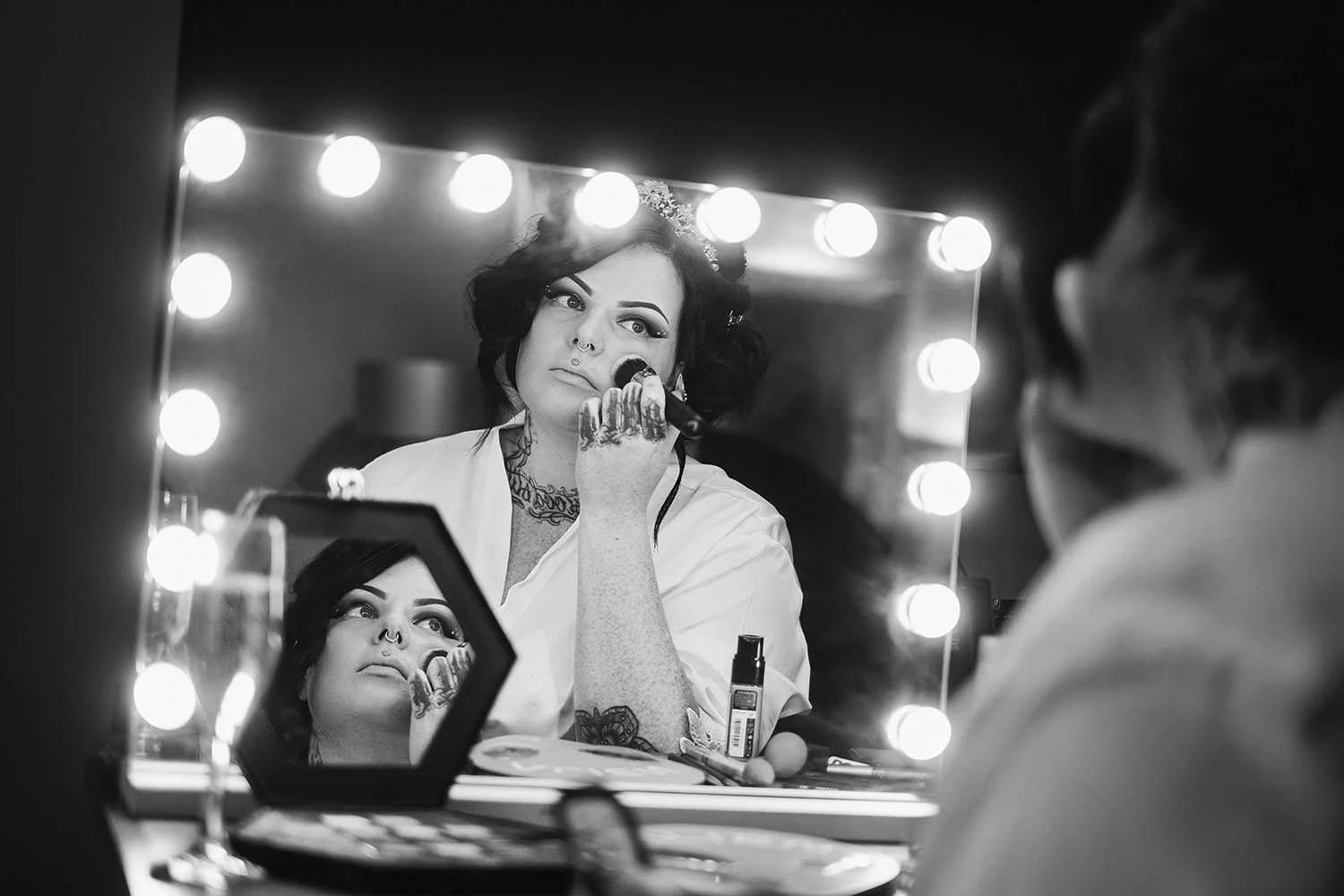 Tattooed bride looking at herself in a mirror ringed with lights while getting ready, in black and white.