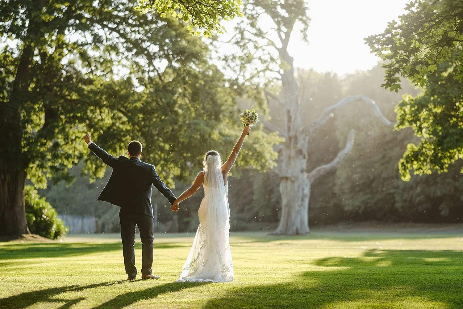 Beamish Hall wedding: bride and groom walking away hand in hand with their arms raised through sunlit parkland.