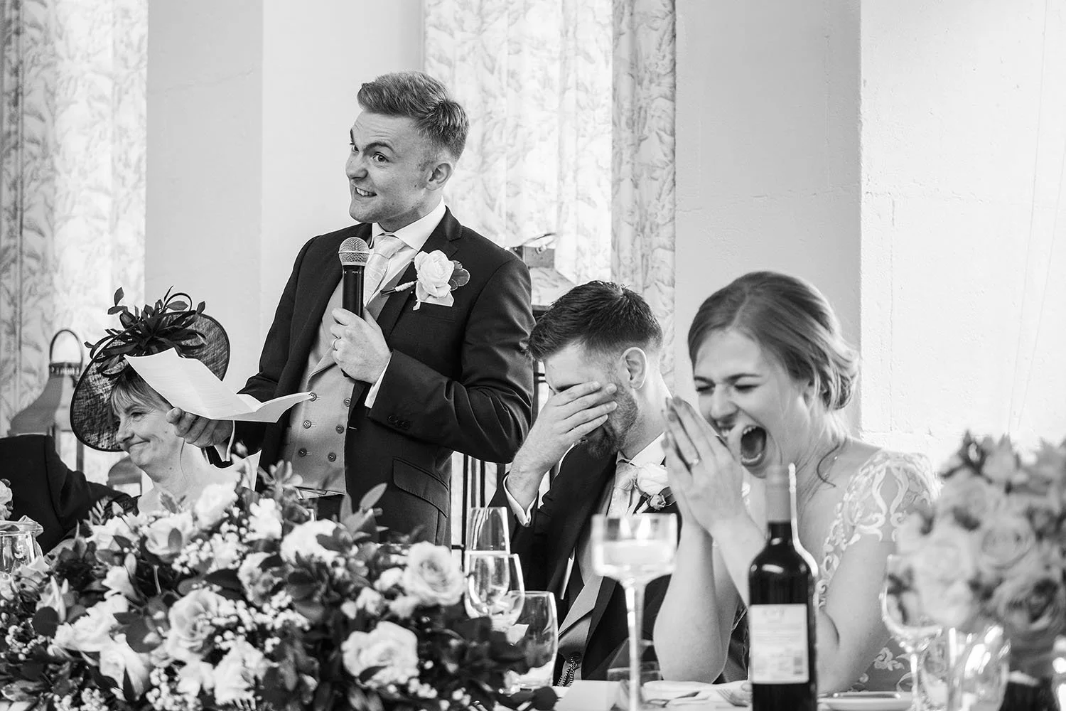 Wynyard Hall wedding: groom giving a speech as the bride laughs with her hands over her face, black and white