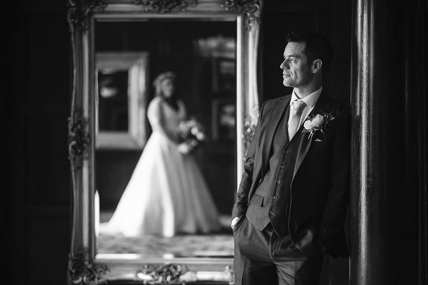 Slaley Hall wedding: groom looking away with the bride reflected in a gilded mirror, in black and white.