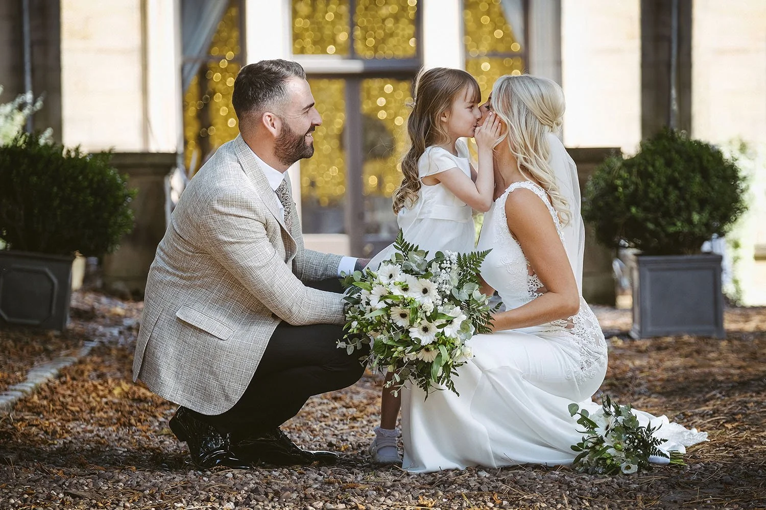 Beamish Hall wedding: flower girl whispering in the bride's ear as the groom kneels beside them in a garden.