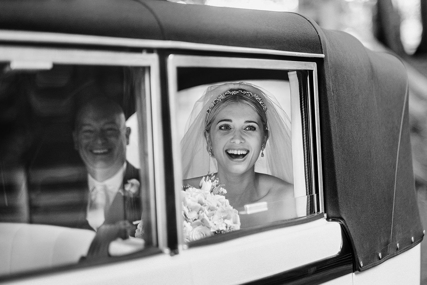 Bride laughing through the window of a vintage wedding car beside her father, in black and white.