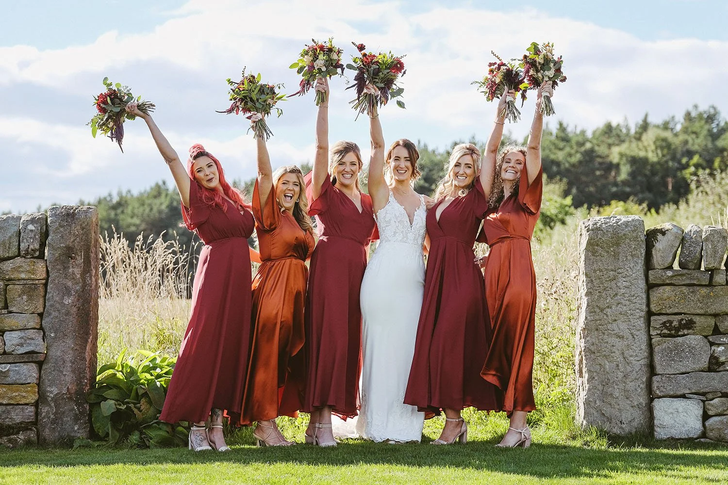 Healey Barn wedding: bride and bridesmaids in red dresses raising their bouquets in the air in front of a stone gateway.