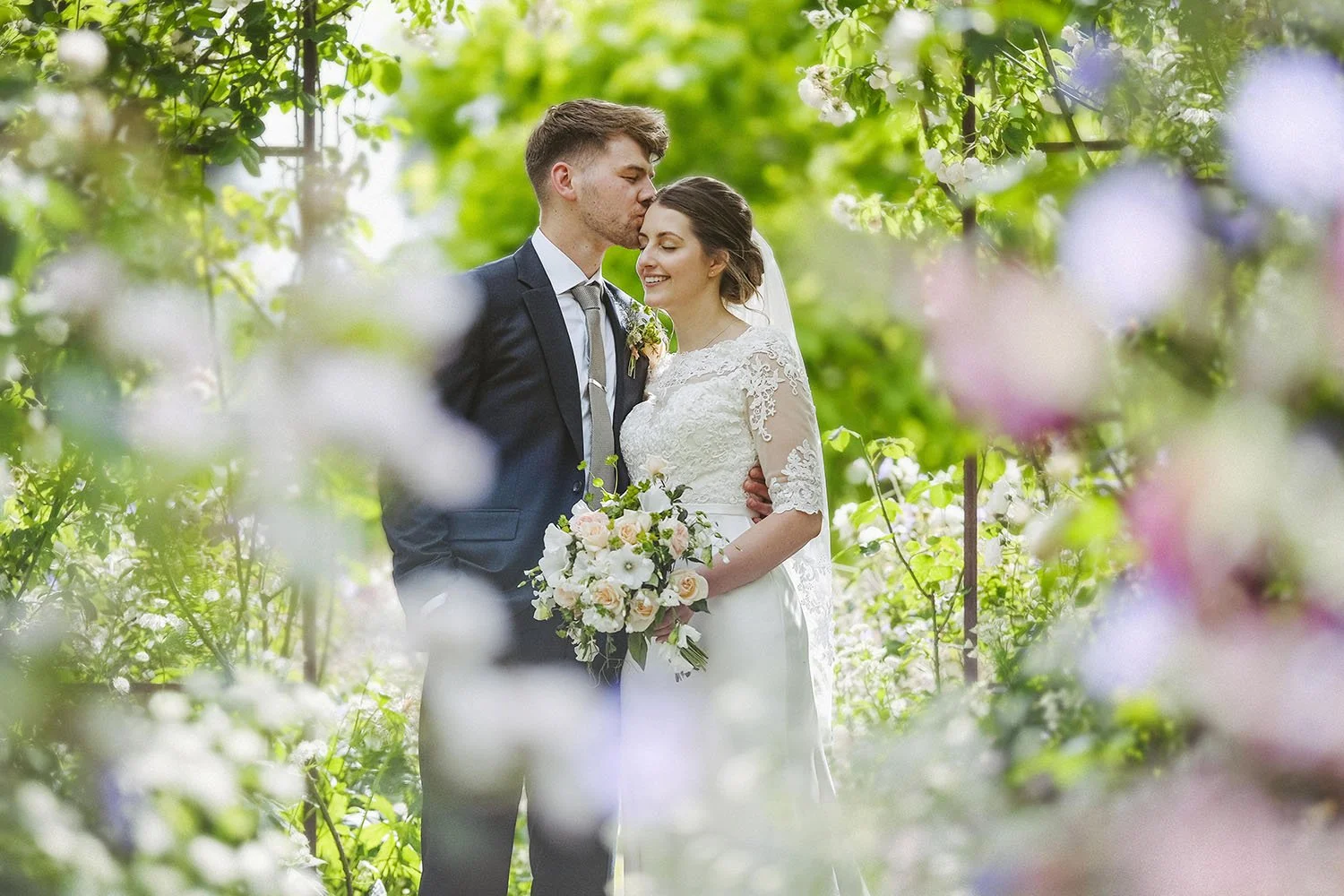 Ellingham Hall wedding: groom kissing the bride's forehead among soft blossom in a garden, the bride holding a bouquet.