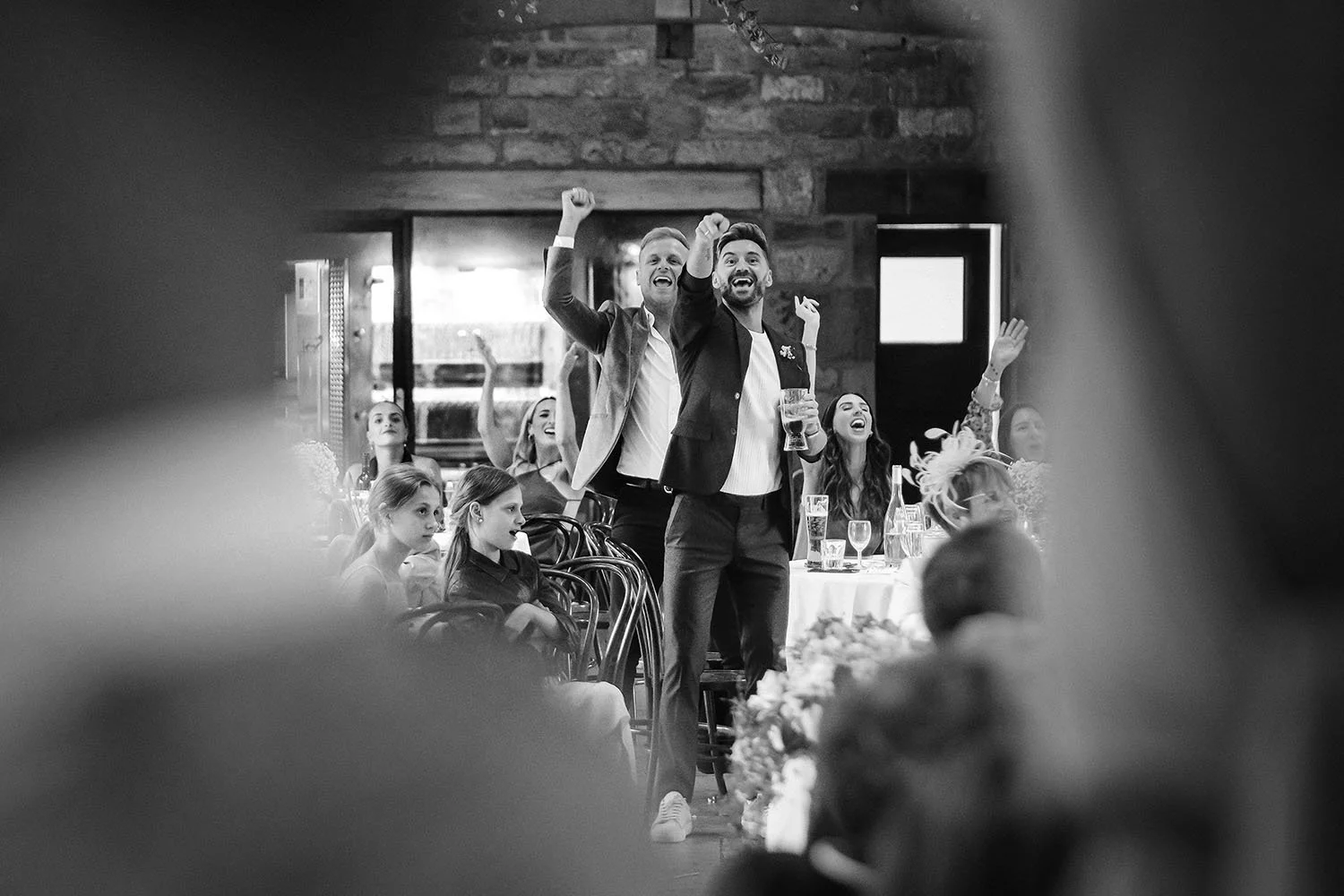 Healey Barn wedding: guests cheering with their arms raised at the reception, seen past a blurred foreground, in black and white.