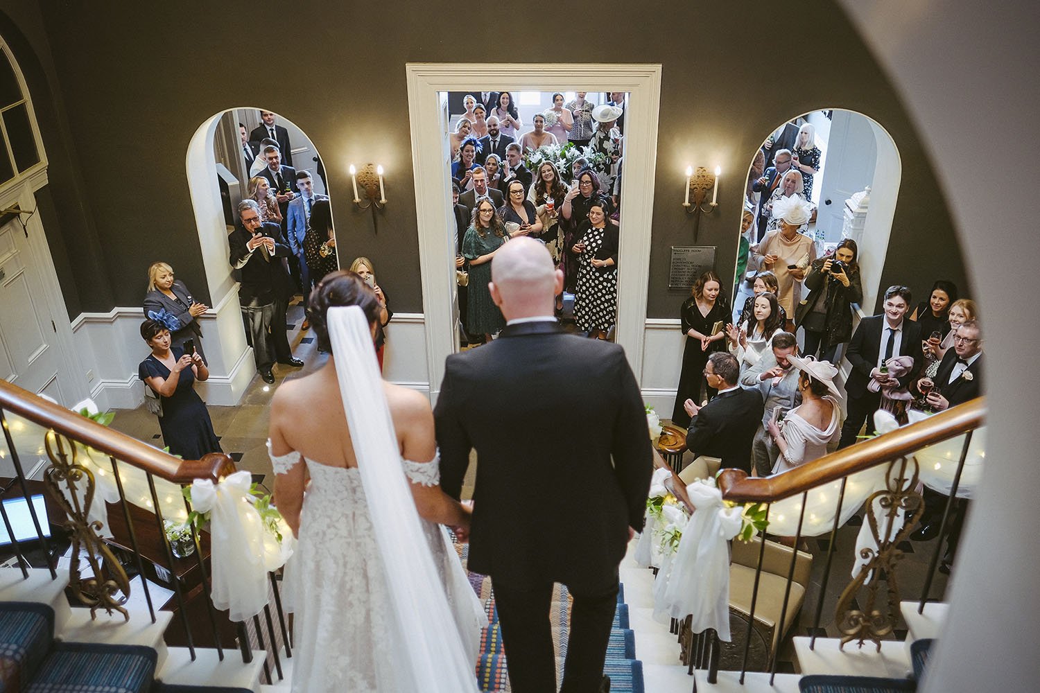 Bride and her father at the top of a staircase seen from above, guests gathered at the door below.