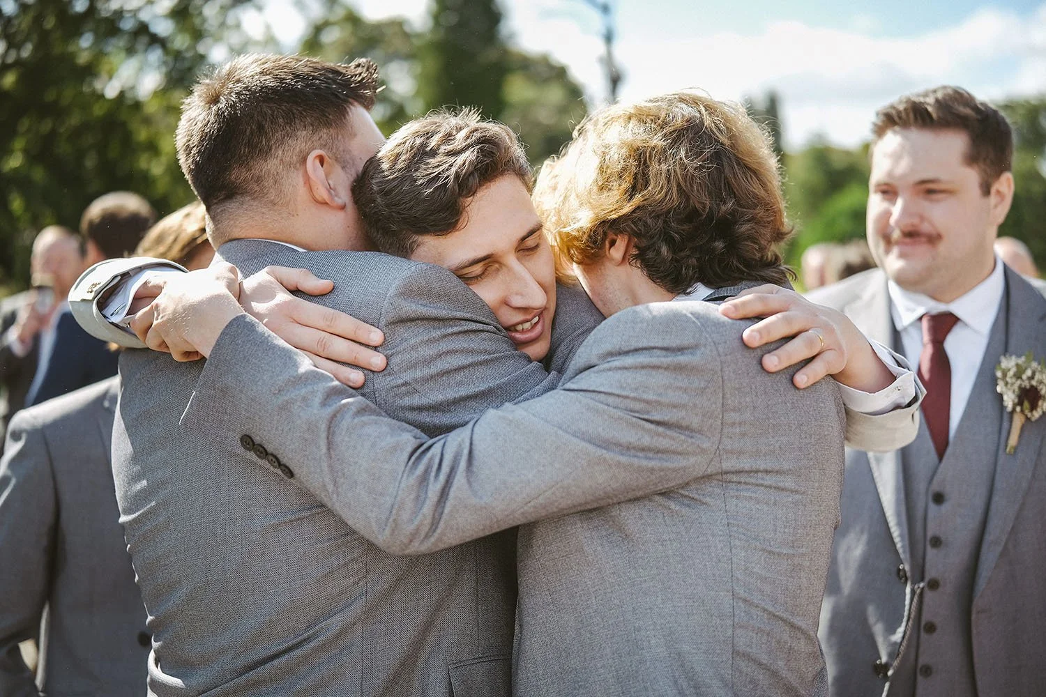 Beamish Hall wedding: groom and two friends in grey suits sharing a three-way hug on a lawn while a bearded guest looks on.