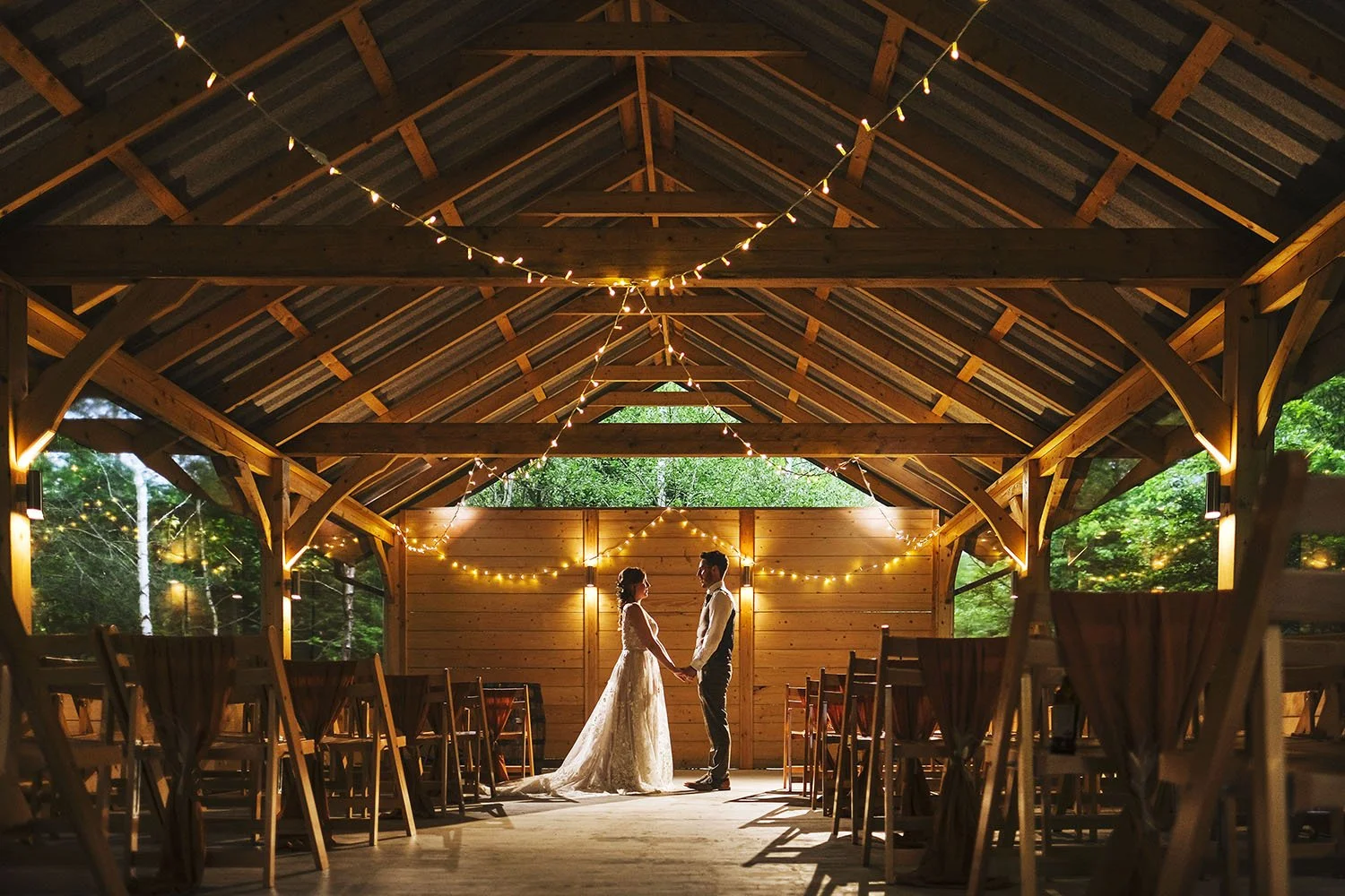 Bradbury Glade wedding: bride and groom holding hands in an open timber-framed barn strung with fairy lights.