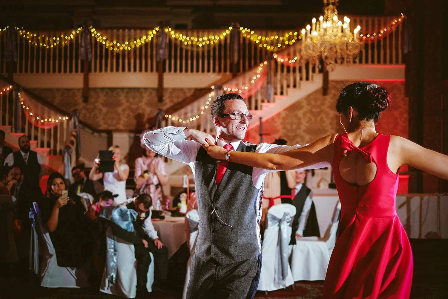 Guisborough Hall wedding: groom dancing with a woman in a red dress beneath a chandelier and fairy lights in a ballroom