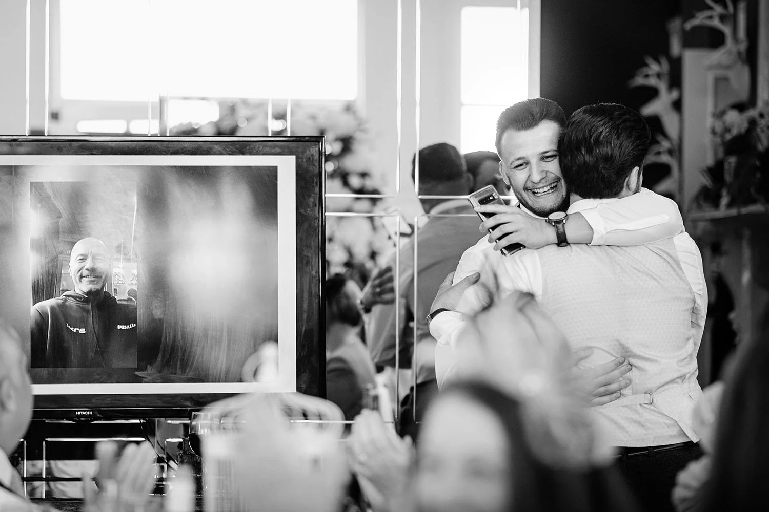 Woodhill Hall wedding: guests hugging and celebrating in front of a big screen at the reception, black and white