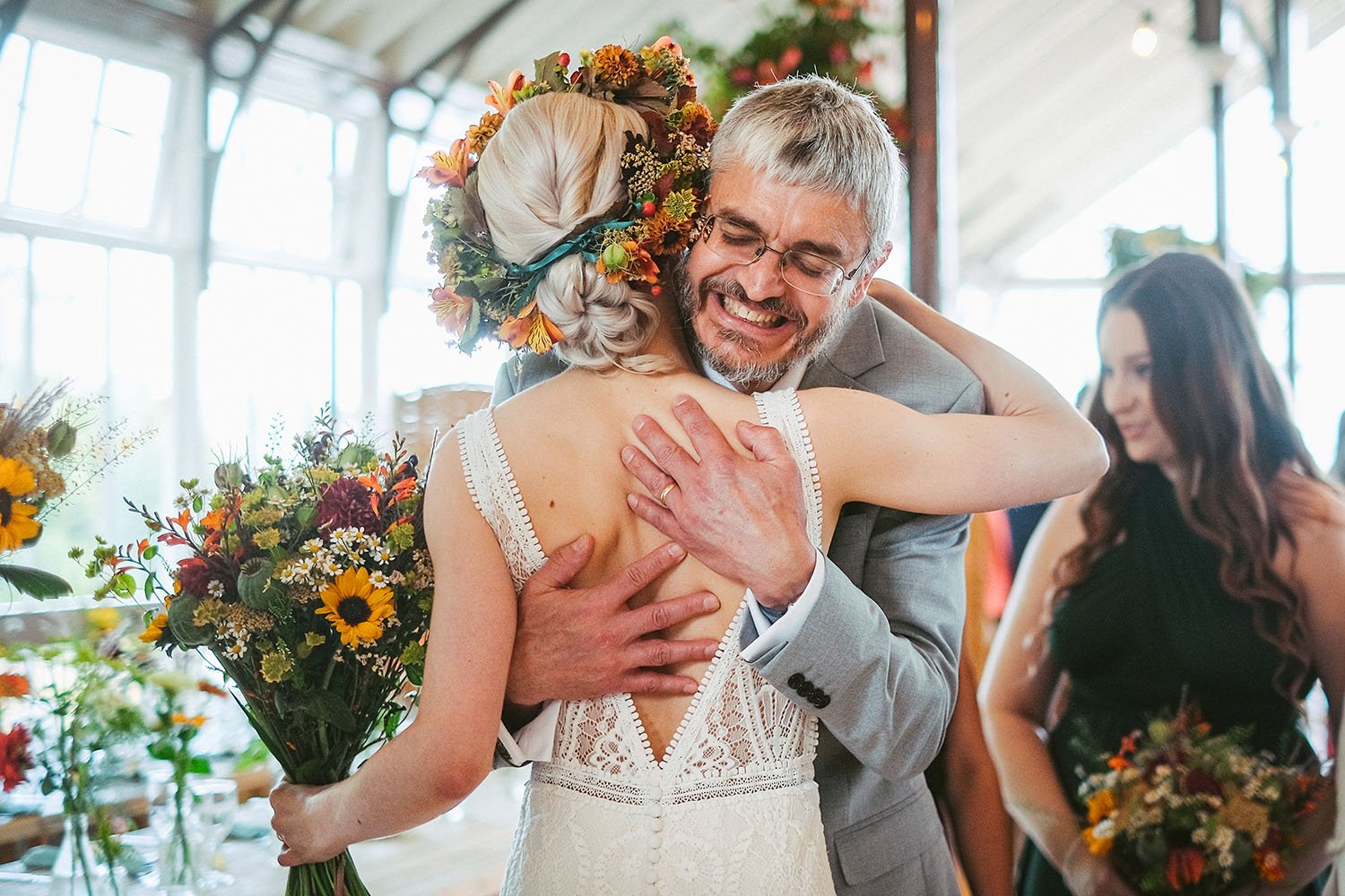Bride wearing a floral crown hugs a smiling man in a gray suit at a wedding reception.