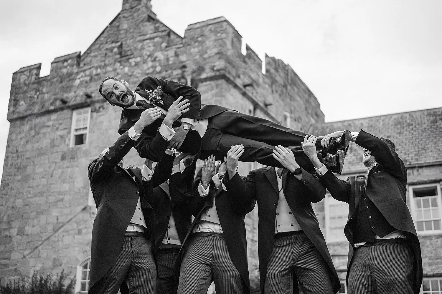 Shortflatt Tower wedding: groomsmen in morning suits lifting the groom into the air in front of a castellated stone tower
