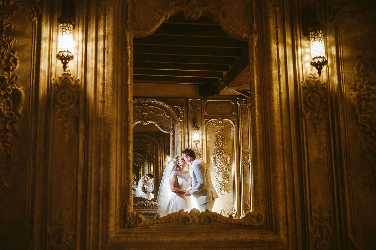 Le Petit Chateau wedding: bride and groom kissing, seen through an ornate gilded mirror frame in a warmly lit room.