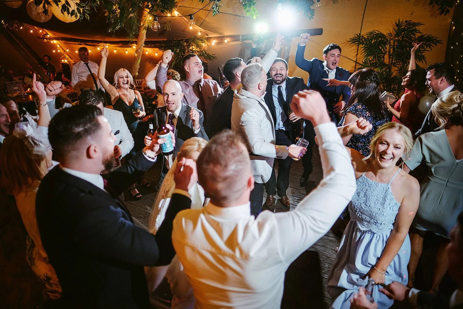 Woodhill Hall wedding: guests dancing with their arms raised at a lively party in a tipi under festoon lights