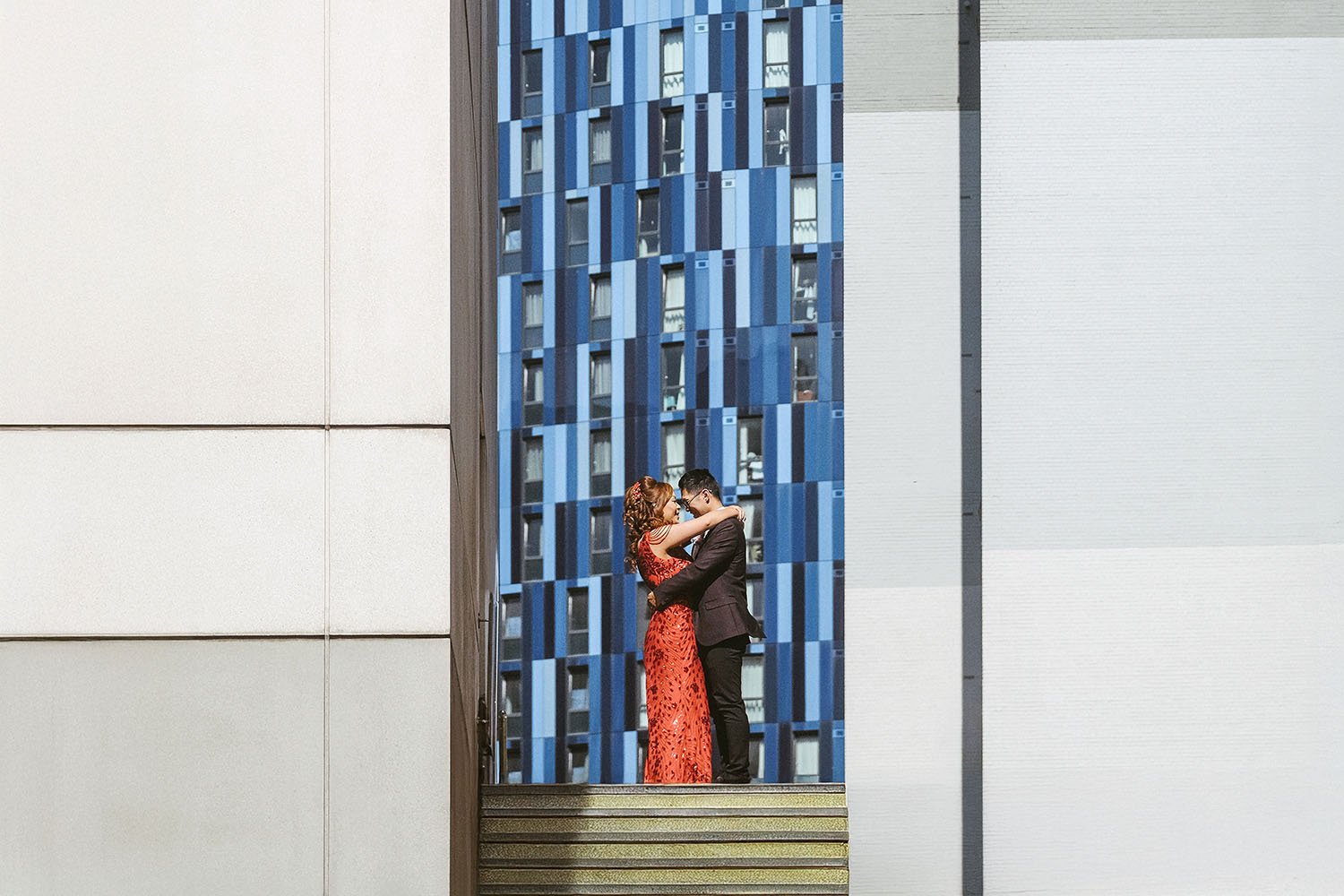 Couple embracing in a doorway in front of a blue patterned building, she in a red dress, framed by white walls.