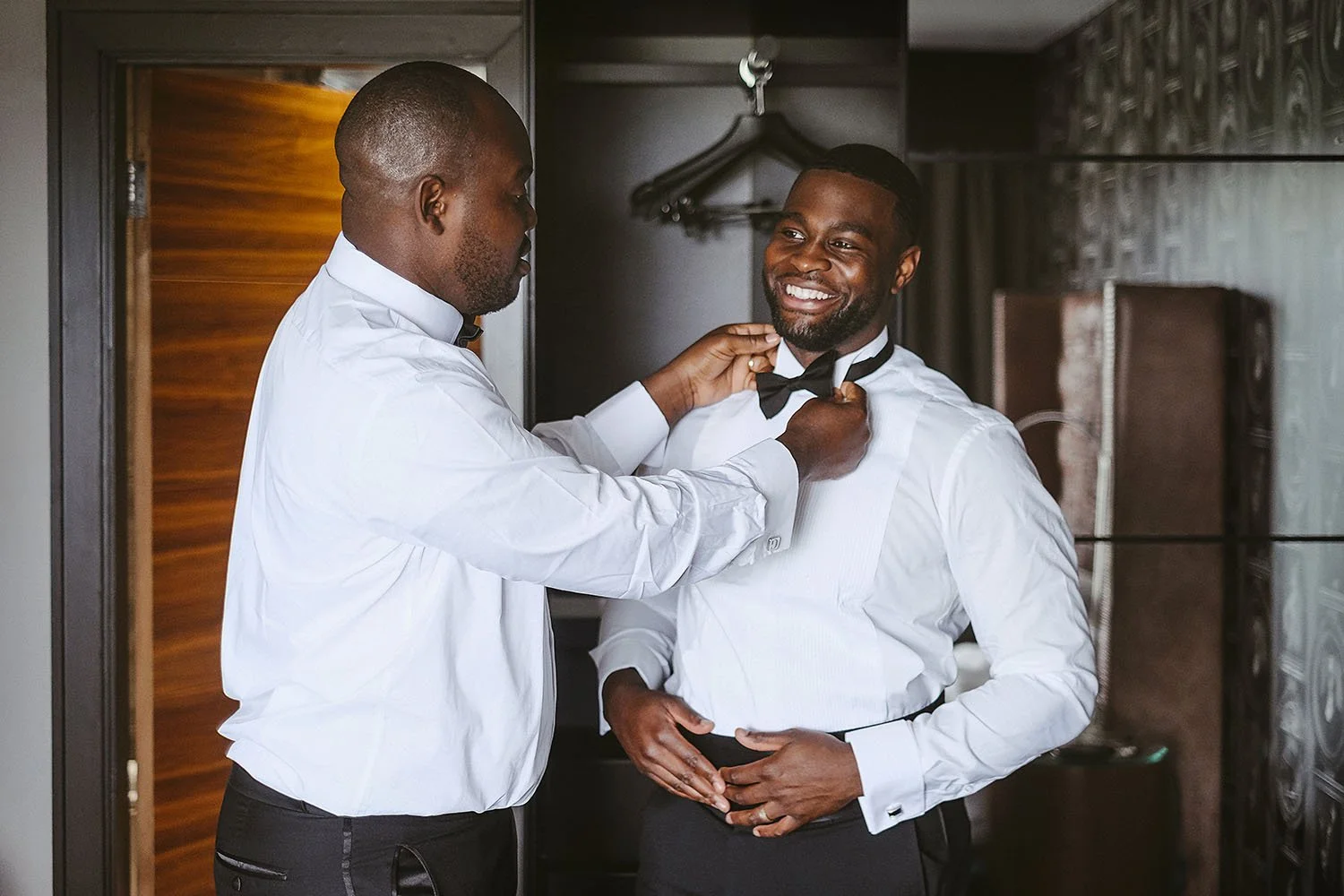 Groom smiling as a friend adjusts his bow tie in a hotel room.