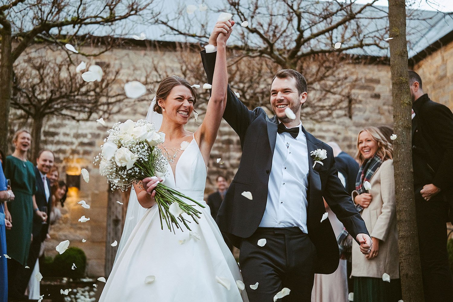 Healey Barn wedding: bride and groom celebrating with arms raised as petal confetti falls in the courtyard.