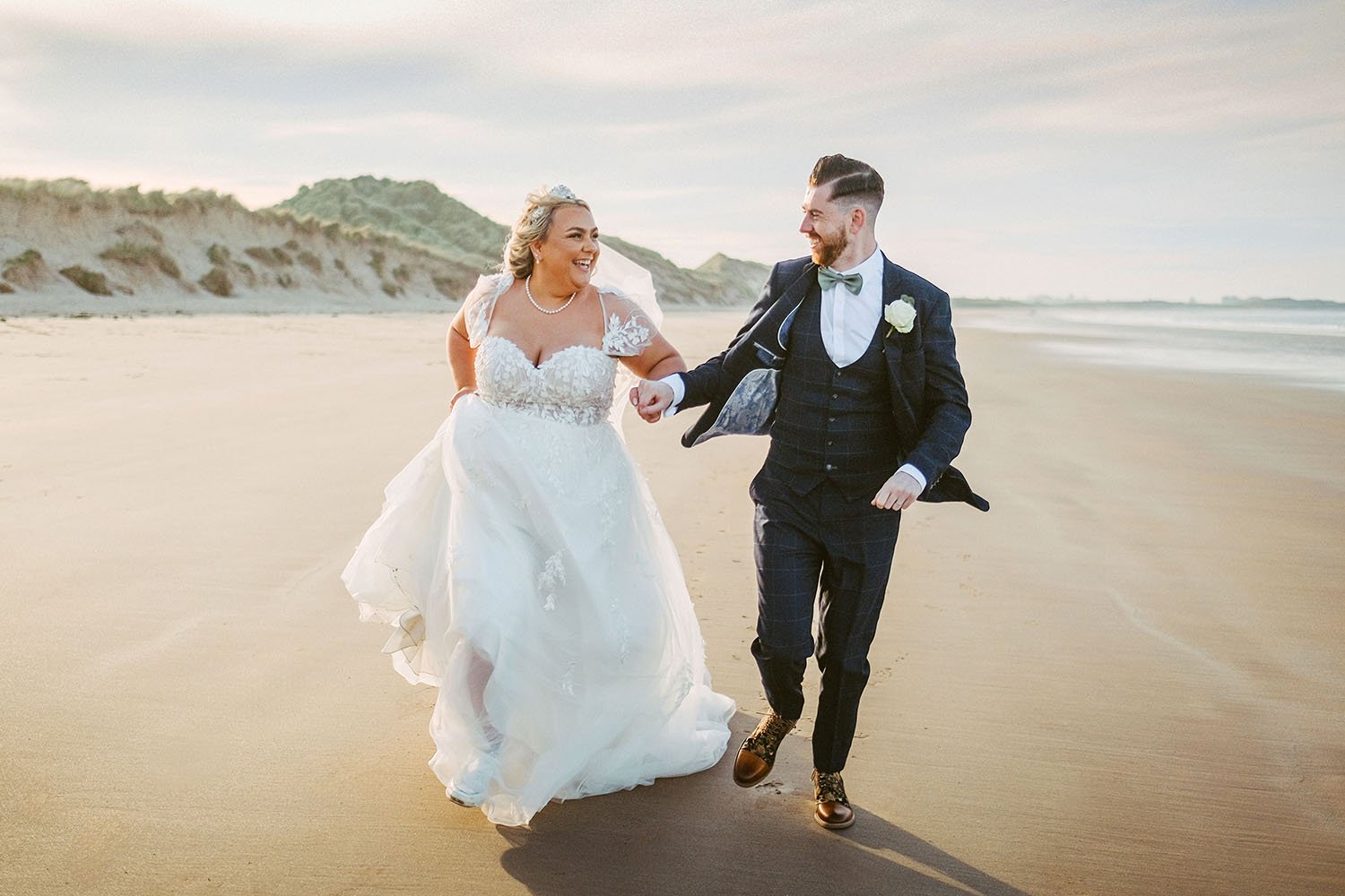 Newton Hall wedding: bride and groom laughing as they run hand in hand along a beach in golden light, dunes behind them.