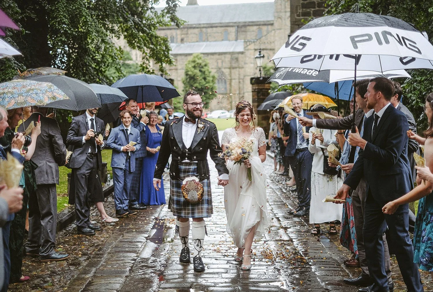 Durham Castle wedding: bride and groom in a kilt walking hand in hand between guests with umbrellas on a wet cobbled path.