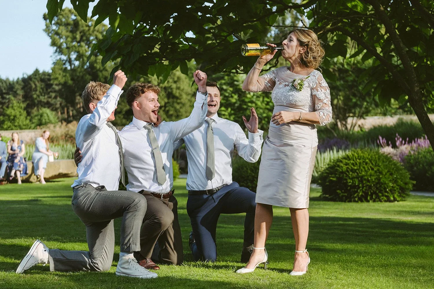 Wedding guests celebrate on a lawn as a woman in a white dress drinks from a bottle and three men cheer.