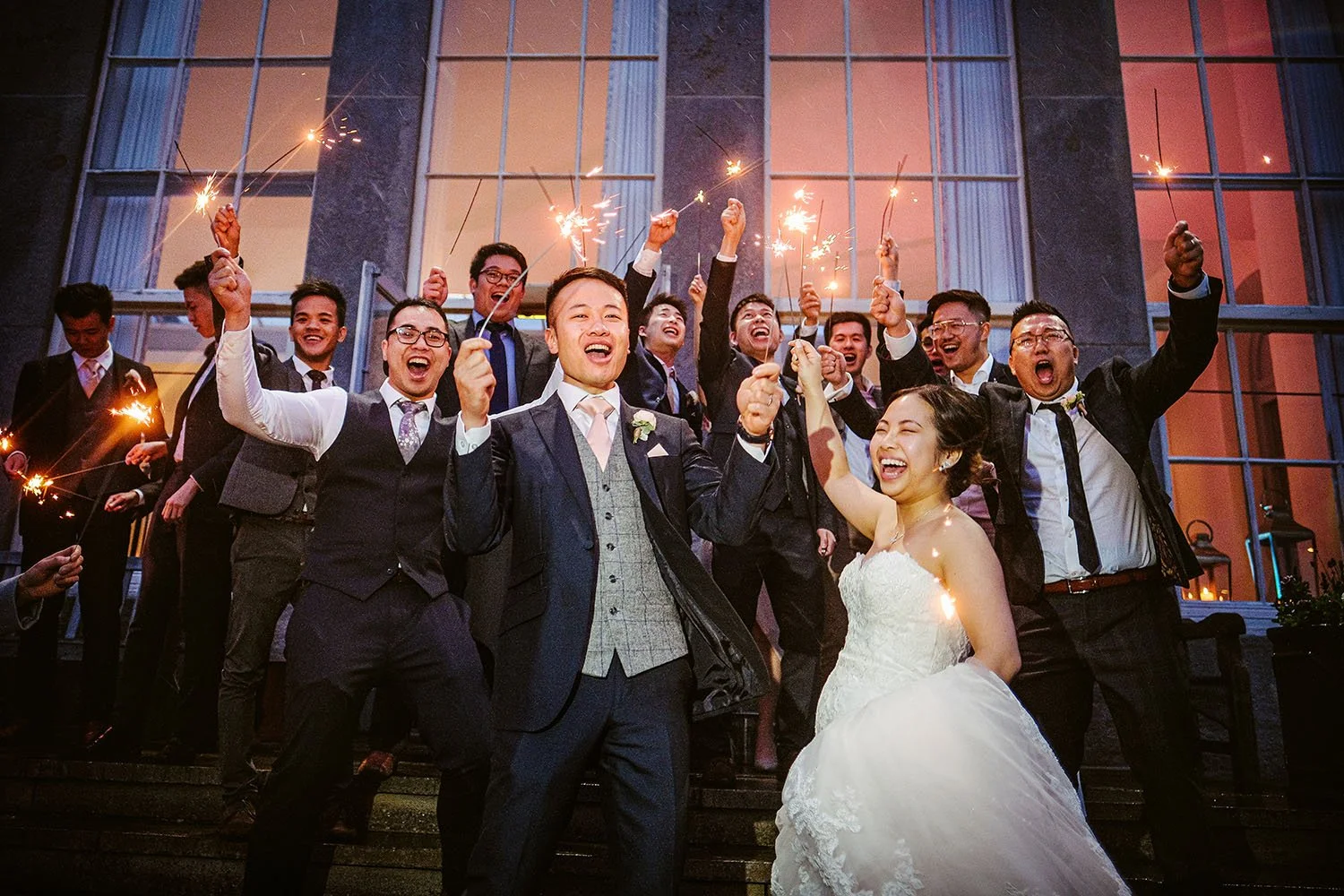 Wynyard Hall wedding: groom, bride and groomsmen cheering with sparklers on stone steps in front of tall windows at dusk