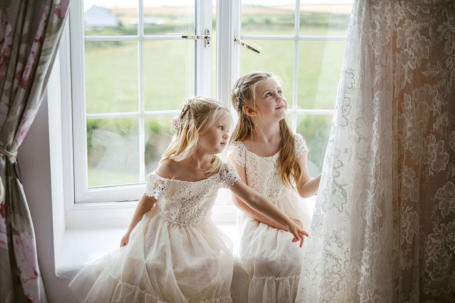 Wynyard Hall wedding: two flower girls in lace dresses sitting on a window sill beside a lace curtain.