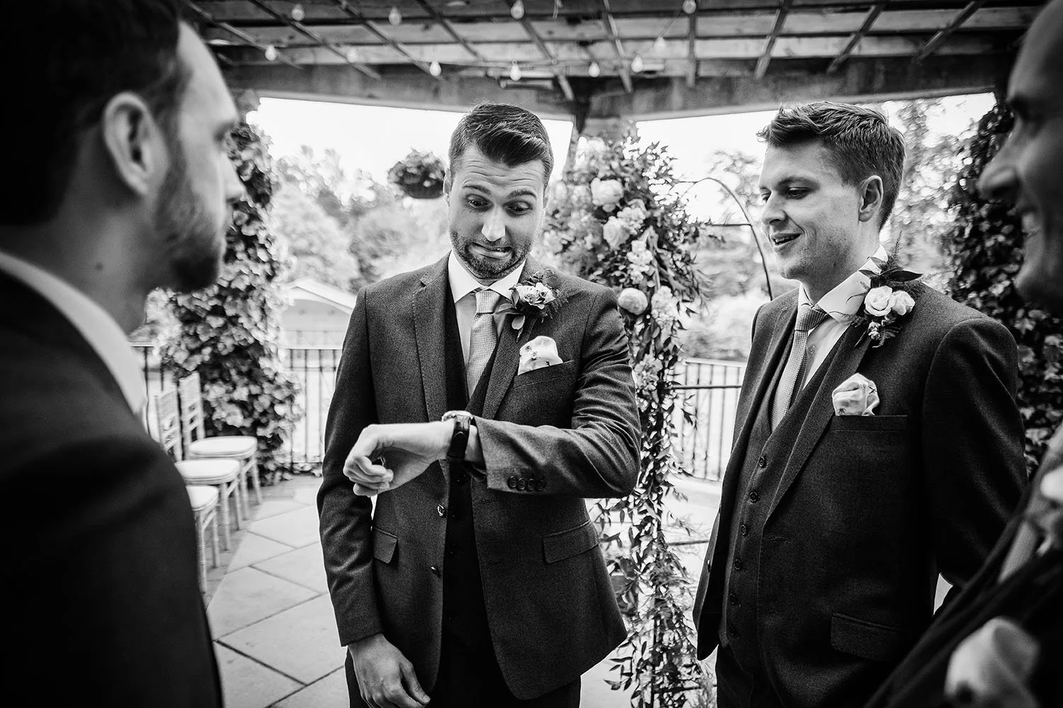 Black-and-white wedding photo of groomsmen in suits and boutonnieres chatting on a covered patio.