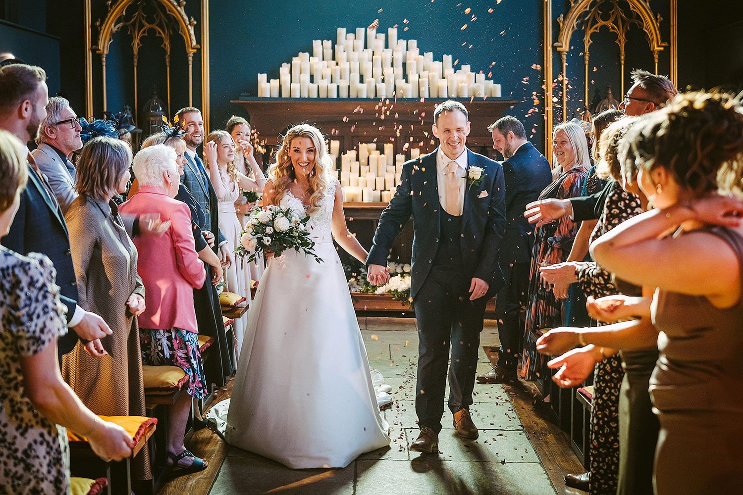 Lartington Hall wedding: bride and groom walking hand in hand up the aisle through petals thrown by guests in a room