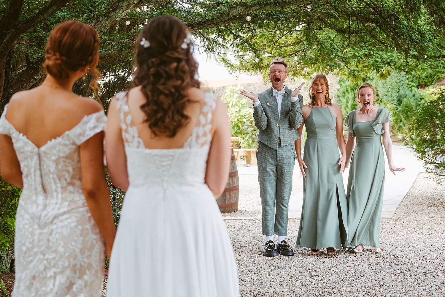 Bride and bridesmaid greet surprised wedding guests outdoors under a tree