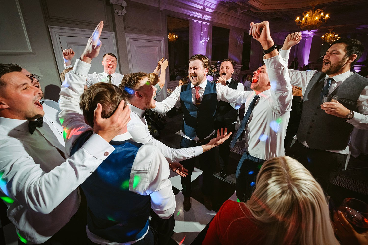 Acklam Hall wedding: groomsmen and guests singing and dancing with their arms raised in a hall with purple lighting.