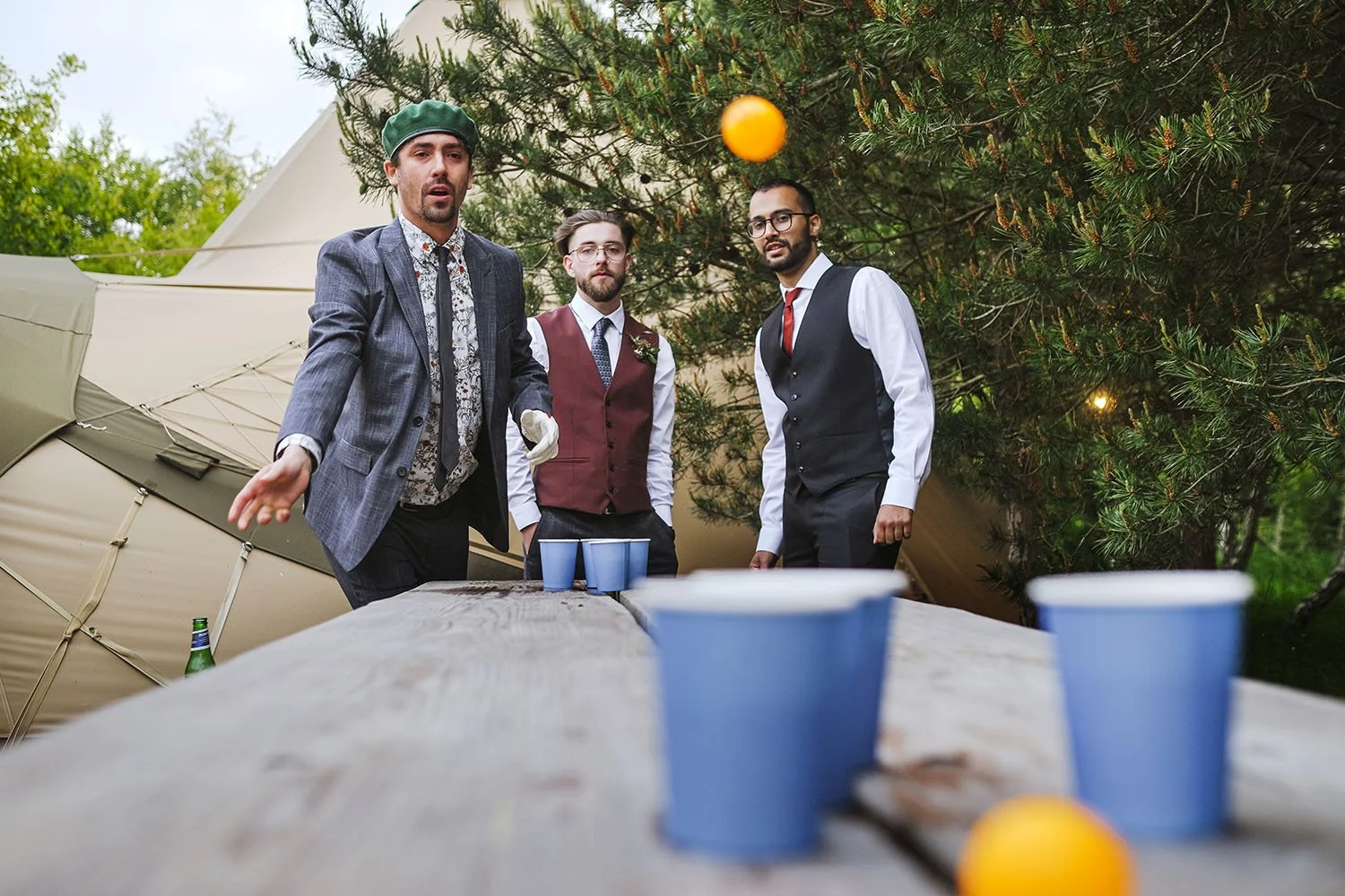 Bradbury Glade wedding: groom and friends playing a ping-pong ball cup game on a wooden table by a tipi.