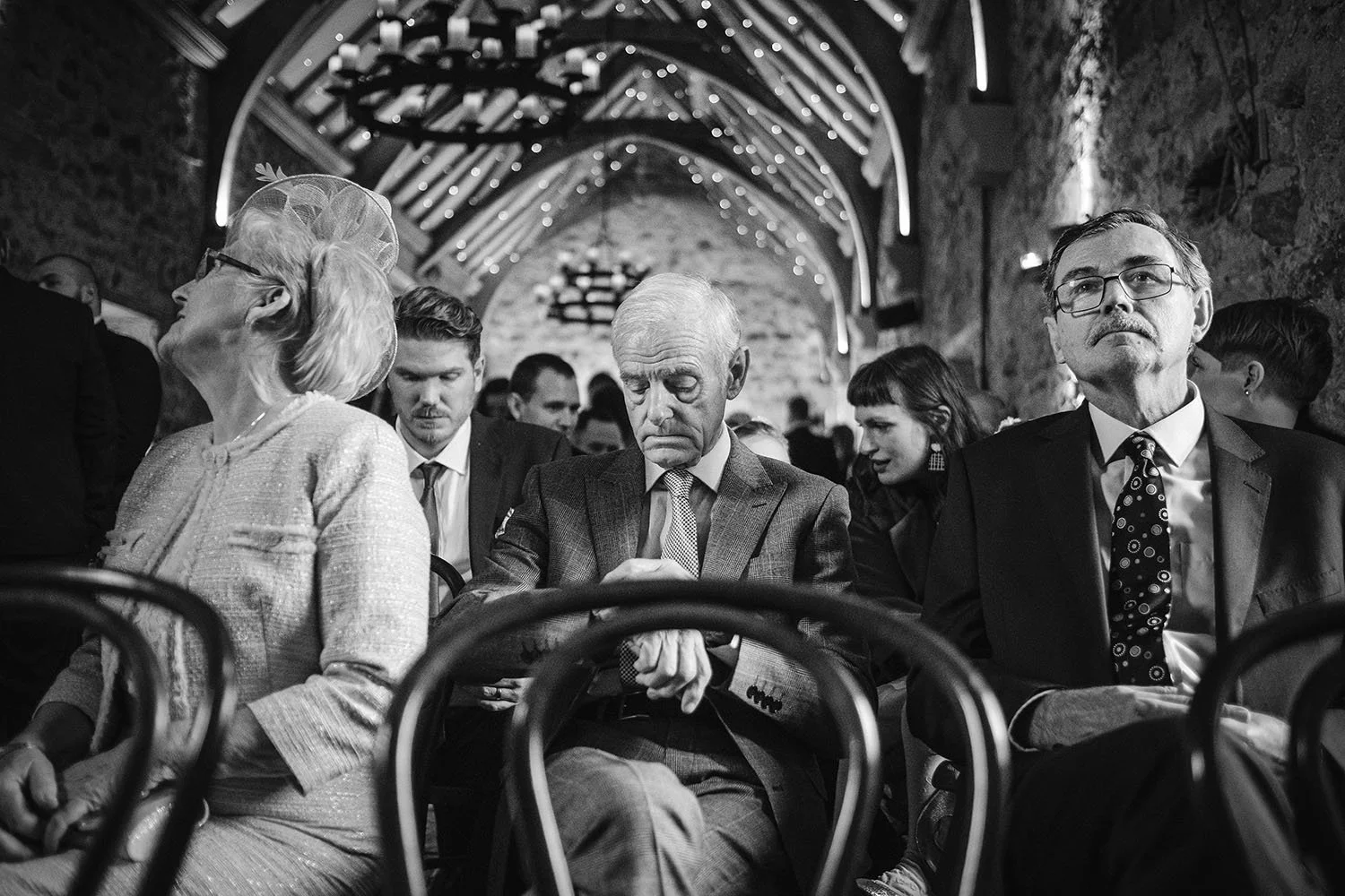 Healey Barn wedding: guests seated on bentwood chairs in a stone barn hung with fairy lights during the ceremony, black and white