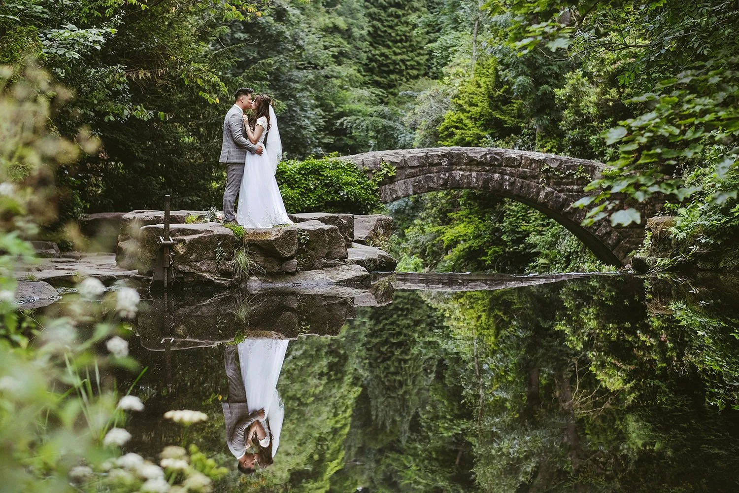 Newlywed couple embracing beside a stone arch bridge in a lush forest, reflected in a calm pond
