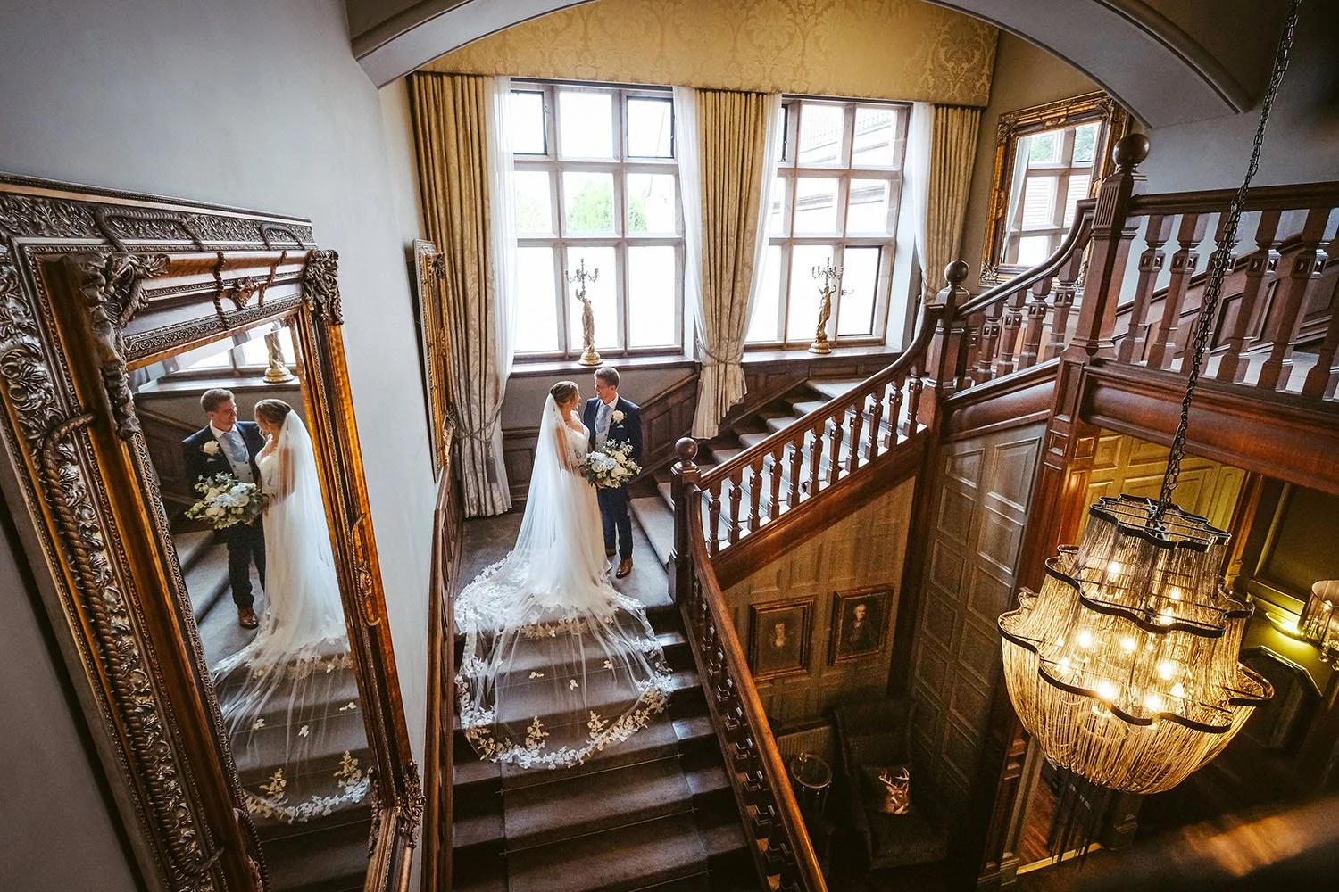 Ellingham Hall wedding: bride and groom on a grand wooden staircase seen from above, reflected in a large gilded mirror