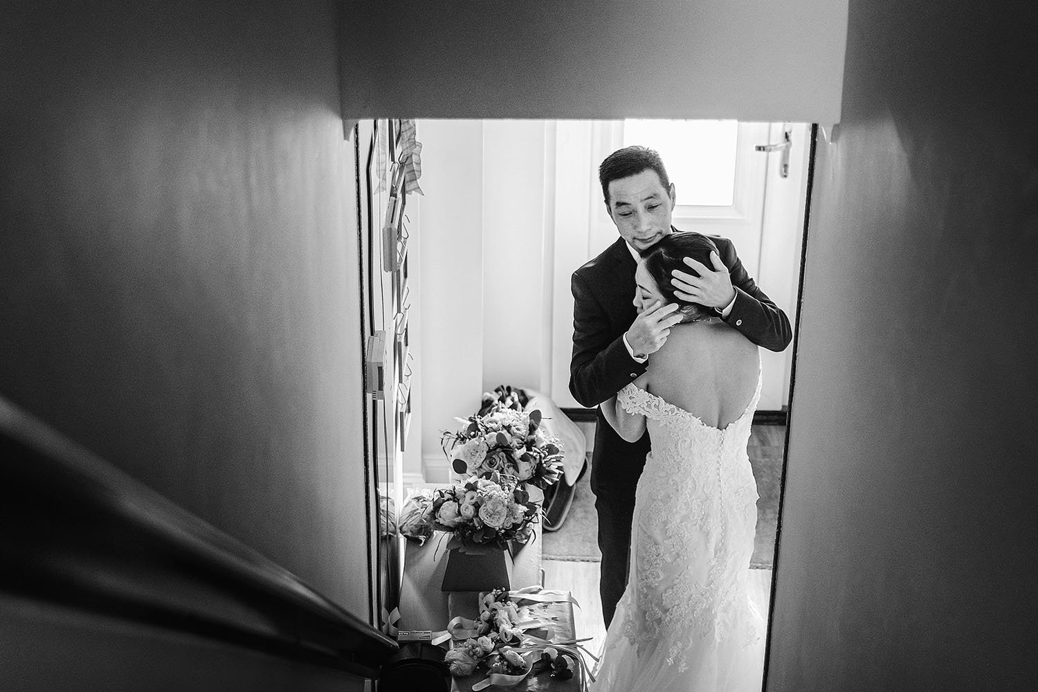 Black-and-white wedding portrait of a groom embracing his bride in a lace wedding dress beside a mirror and floral bouquets
