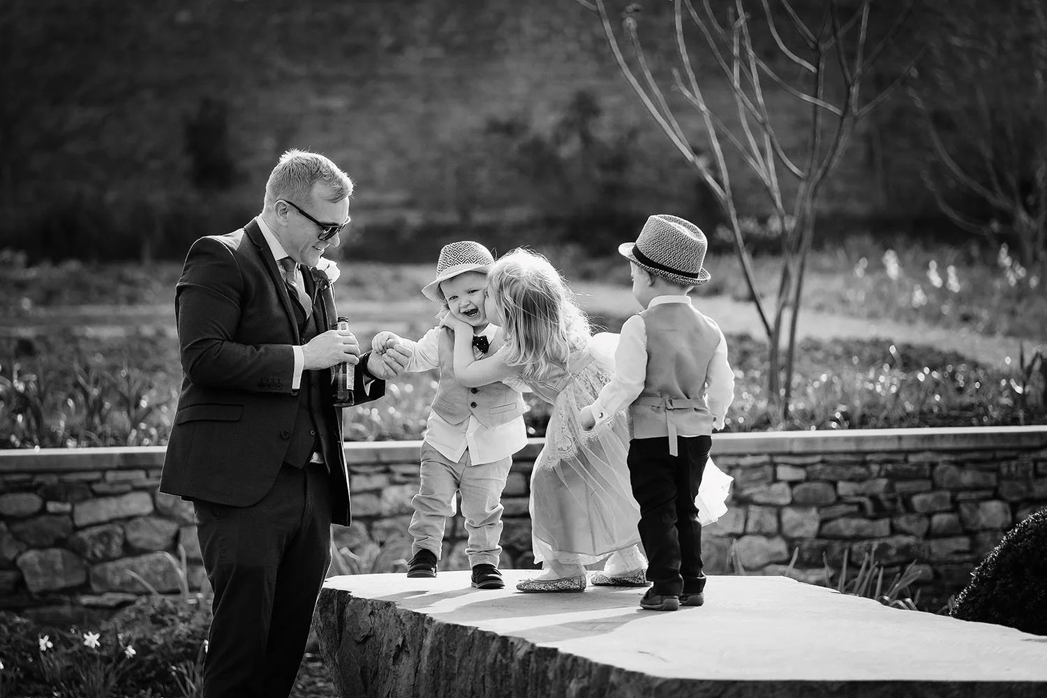 The Fig House wedding: groom with a toddler, a flower girl and a boy in a hat on a stone ledge, black and white