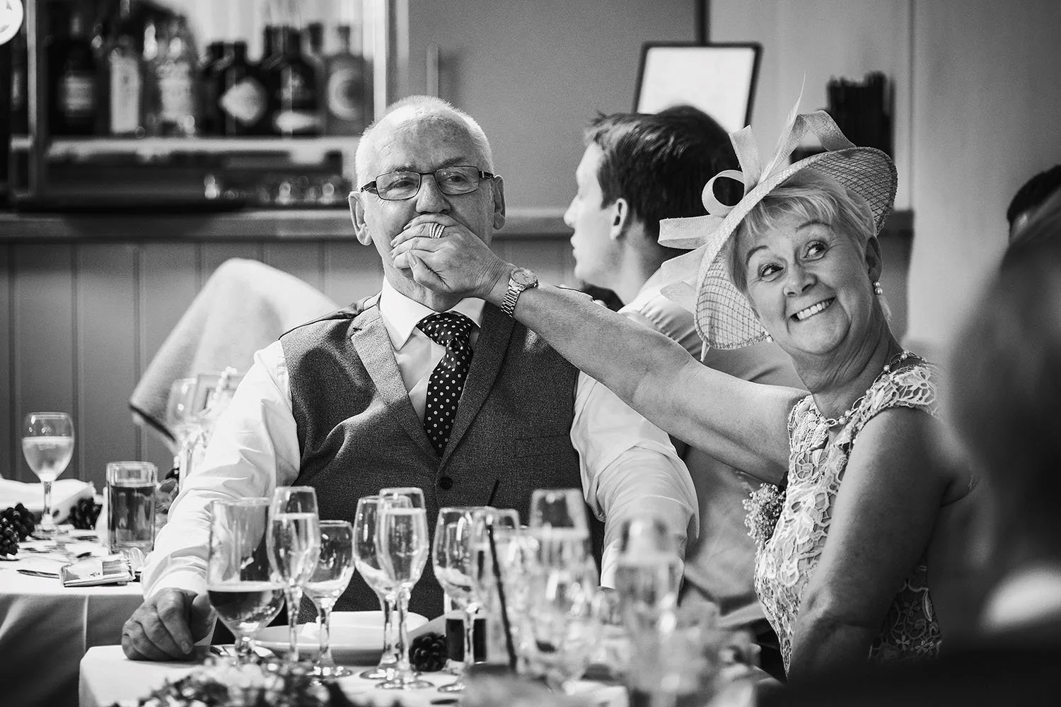 The Parlour at Blagdon wedding: woman in a fascinator playfully covering a man's mouth at the wedding breakfast, black and white
