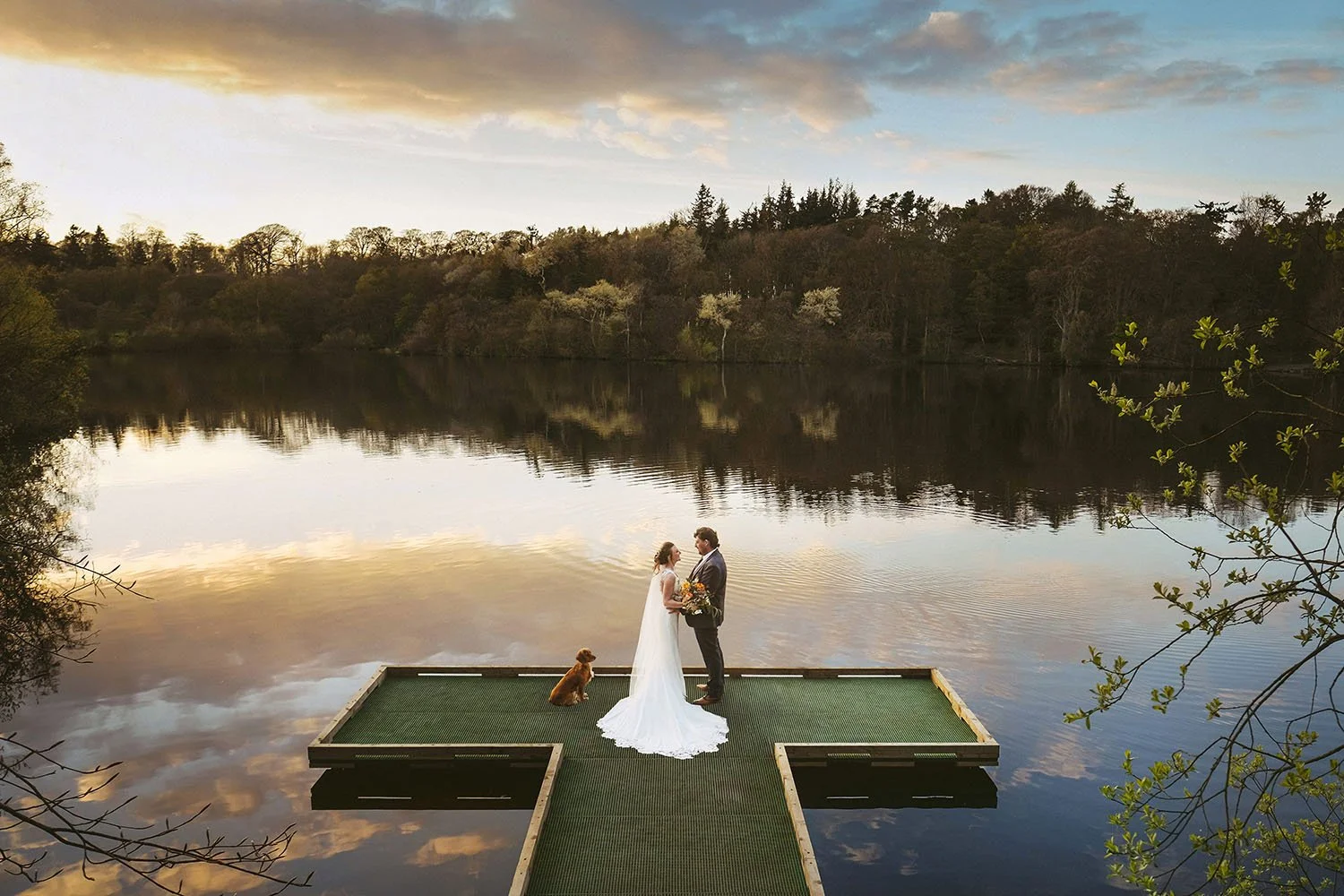 Shortflatt Tower wedding: bride and groom embracing on a wooden jetty with their dog, reflected in still lake water at sunset.