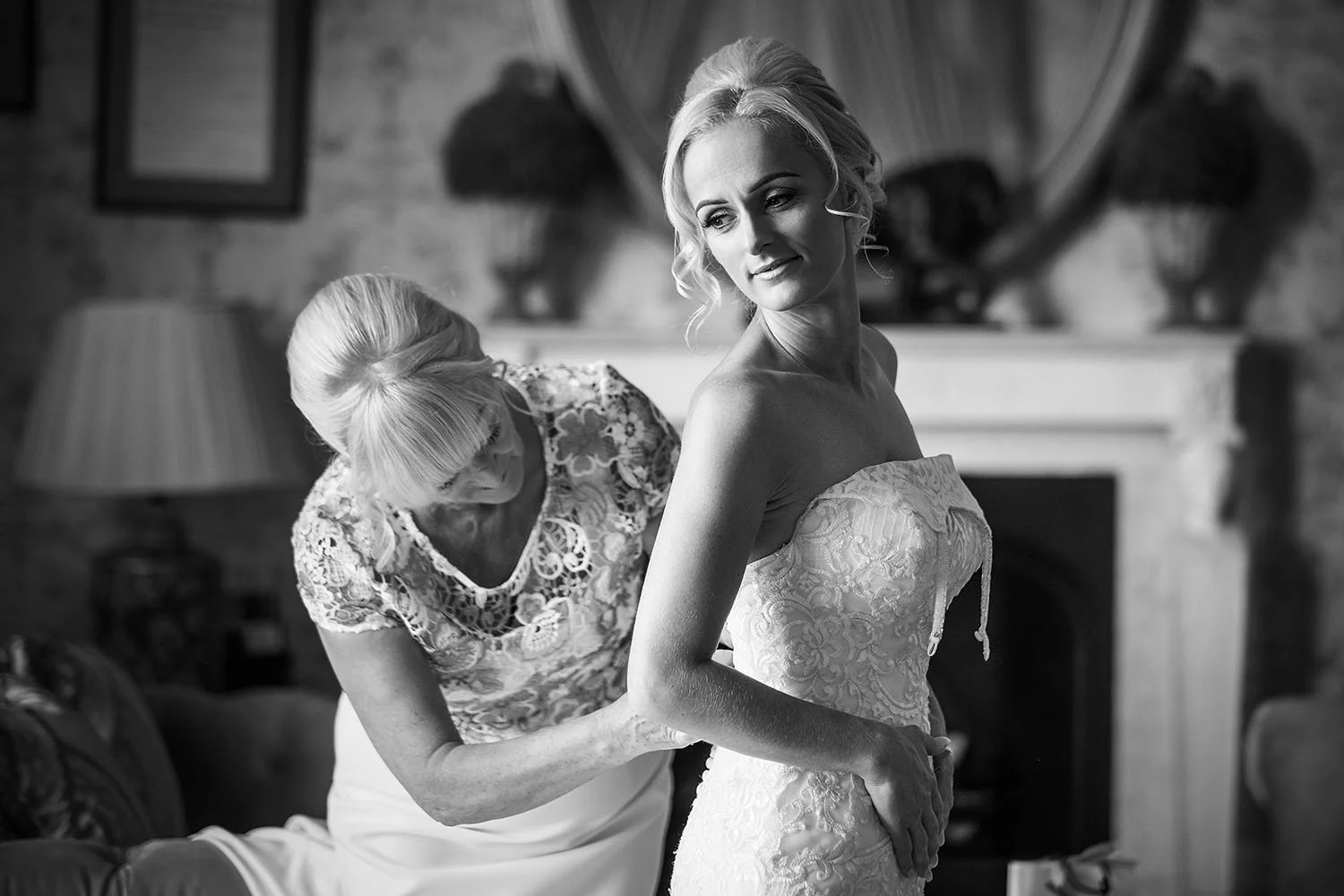Mother helping the bride into her strapless lace dress in front of a fireplace, black and white