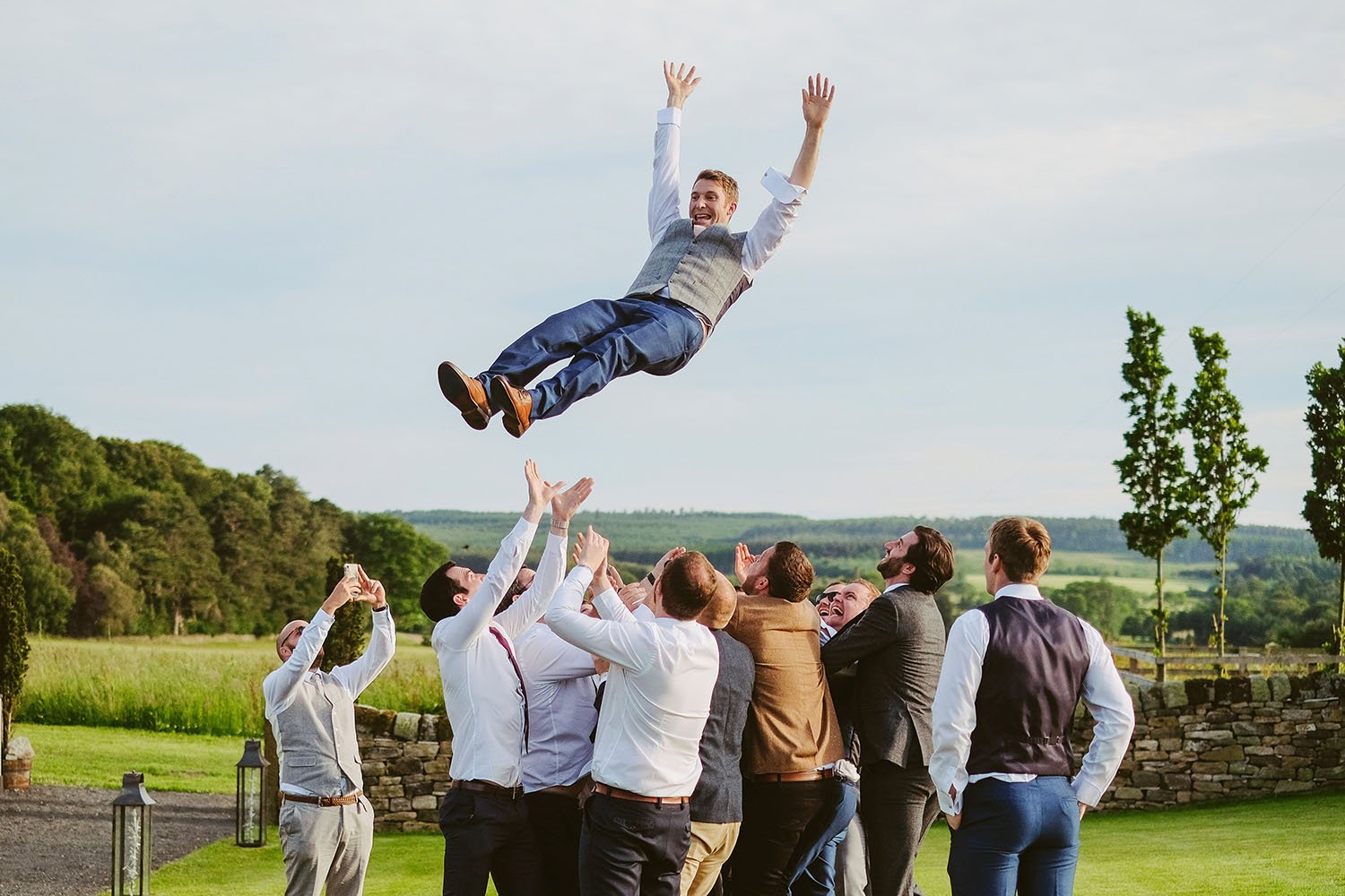Healey Barn wedding: groomsmen tossing the groom high into the air on a lawn with countryside behind