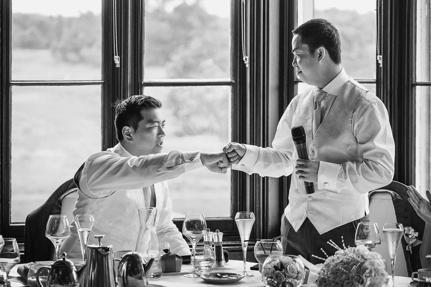 Matfen Hall wedding: groom and best man fist bumping during a speech beside tall windows, black and white